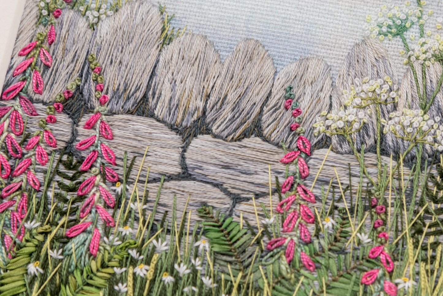 close up of a hand embroidery artwork of a dry stone wall with pink foxgloves, ferns, daisy and grasses in front