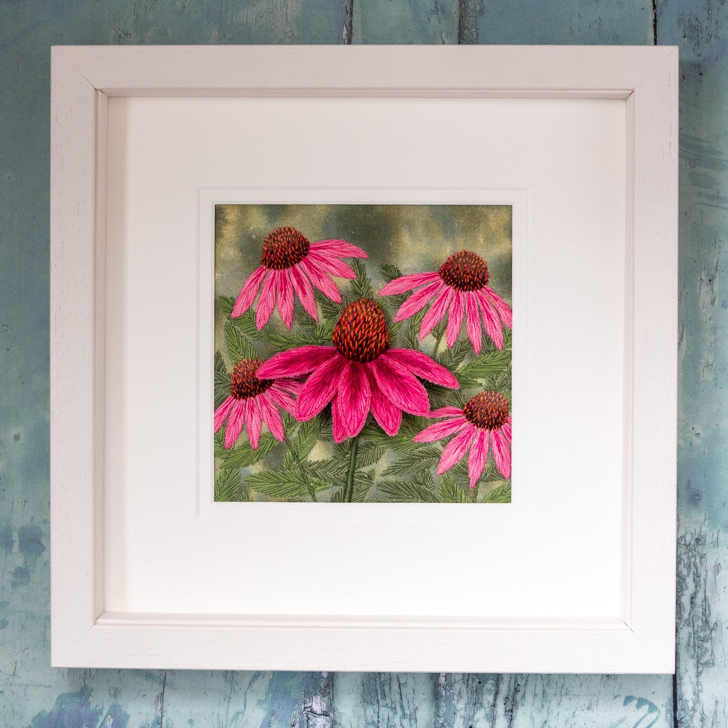 this framed artwork by Jo butcher is a pink echinacea stump work. A large pink 3D cone flower protrudes from the background of other sewn pink cone flowers and their foliage. 
